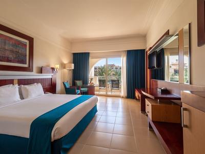 Světý hotelový pokoj s velkou postelí, psacím stolem, televizí a balkonem s výhledem na město.