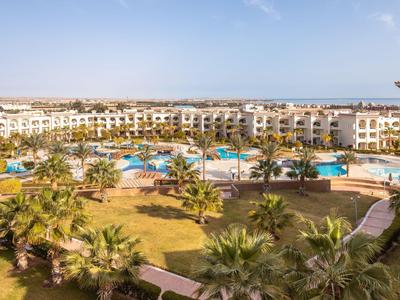 Velký hotelový komplex s bazénem, palmami a jasně modrou oblohou za slunečného dne.