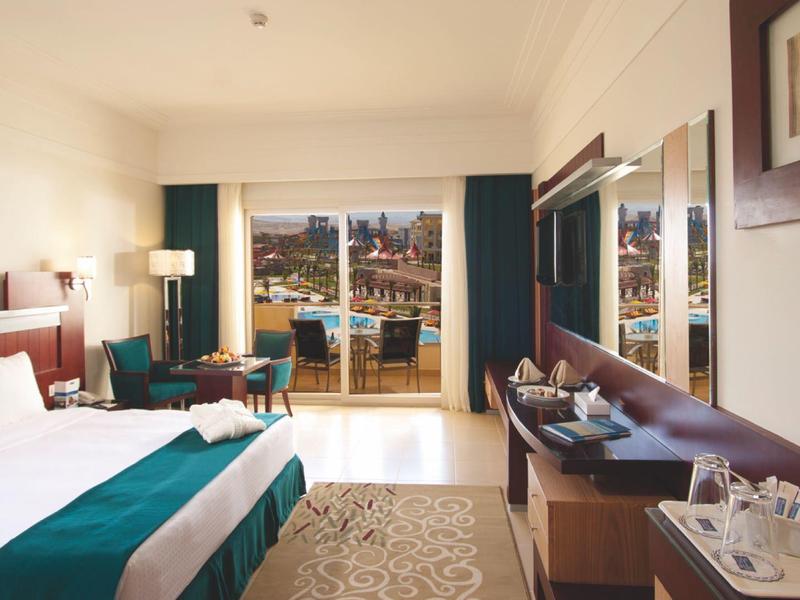 Bright hotel room with double bed, seating area, and balcony overlooking the sea.