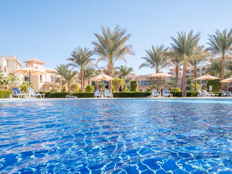 View of a pool with palm trees and sun loungers in front of buildings under clear sky.
