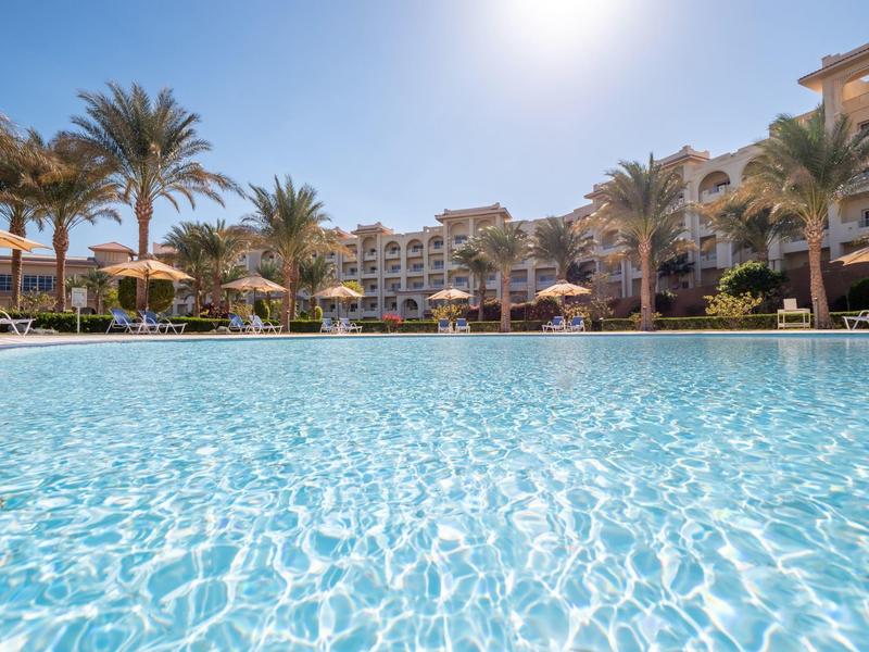 Large outdoor pool in front of a multi-story hotel building under a bright sky with palm trees.