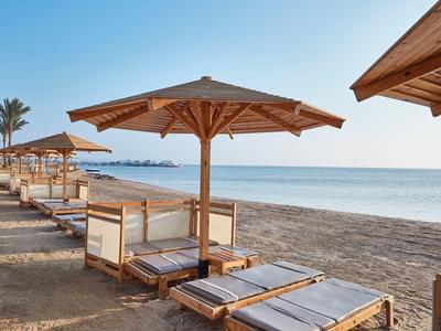 Strand mit Holzliegen und Sonnenschirmen, blauer Himmel und ruhiges Meer im Hintergrund.