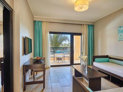 Helles Wohnzimmer mit Sofa, Tisch, Schreibtisch, TV und Balkon mit Blick auf Pool und Palmen.