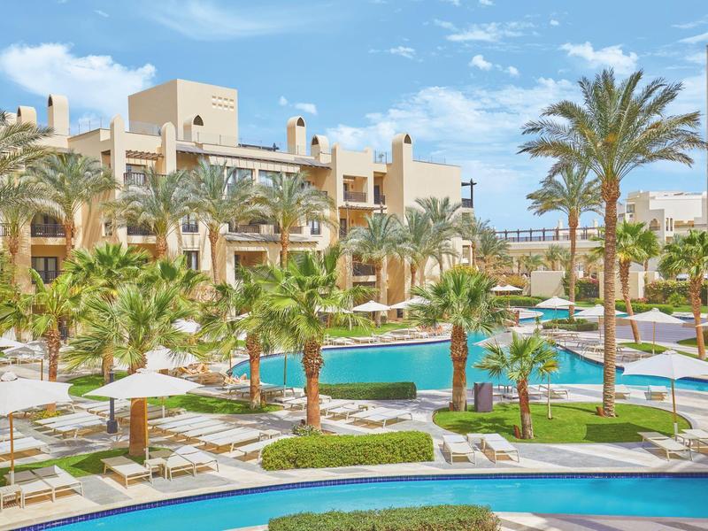 Großer Poolbereich mit Palmen, Sonnenschirmen und modernen Gebäuden unter blauem Himmel.