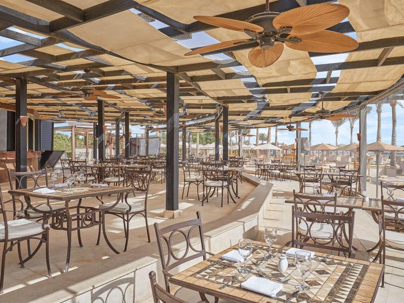 Große offene Terrasse mit vielen Tischen und Stühlen, Holzdeckenventilatoren und Blick auf das Meer.