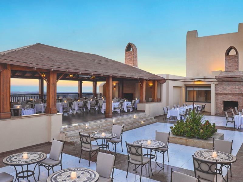 Offene Terrasse mit Tischen, Stühlen, Kamin und Meerblick bei klarem Himmel am Abend.