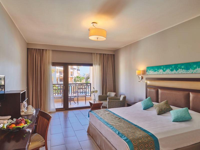 Helles Hotelzimmer mit Doppelbett, Balkon, Fernseher, Schreibtisch und bequemen Stühlen.