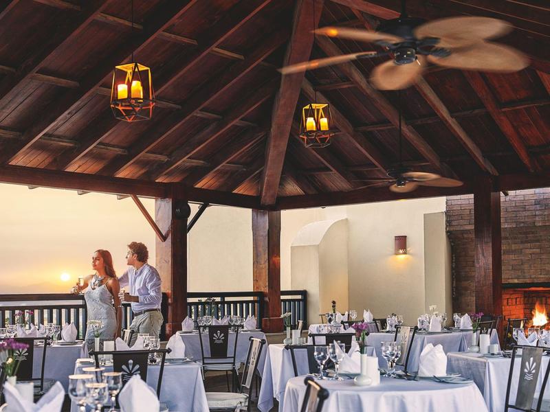 Offenes Restaurant mit Holzdach, gedeckten Tischen und Gästen bei Sonnenuntergang am Meer.