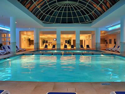 Hallenbad mit blauem Wasser, Liegestühlen und großem, gewölbtem Glasdach.