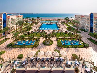 Ampia area piscina dell'hotel con lettini, palme e vista sul mare sullo sfondo.