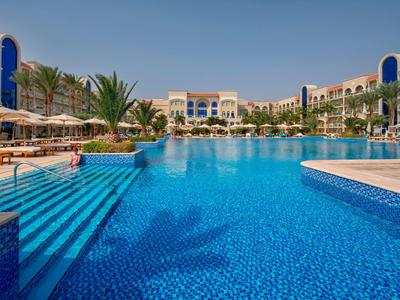 Grande piscina all'aperto con scale, ombrelloni e edifici dell'hotel sullo sfondo sotto un cielo azzurro.