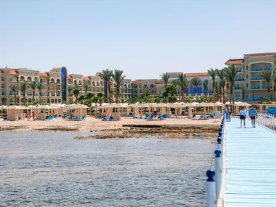 Una spiaggia con ombrelloni, lettini e palme davanti a un complesso alberghiero sotto un cielo limpido.