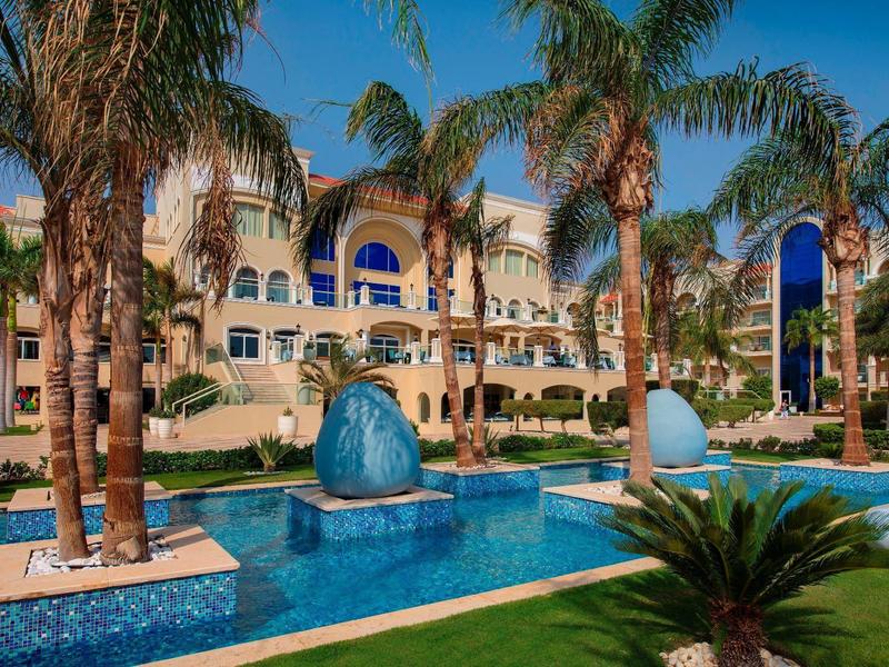 Großes Hotel mit Palmen, blauem Pool und Springbrunnen bei klarem Himmel.