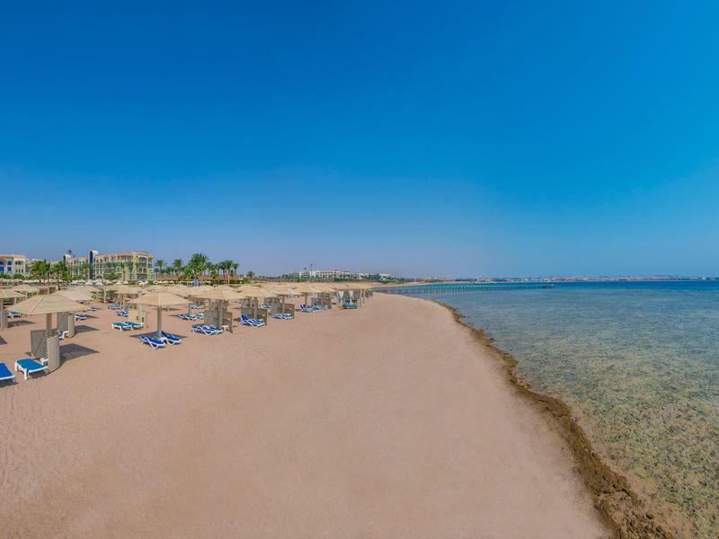 Breiter Sandstrand mit Liegestühlen und klarem, ruhigem Meer unter blauem Himmel.