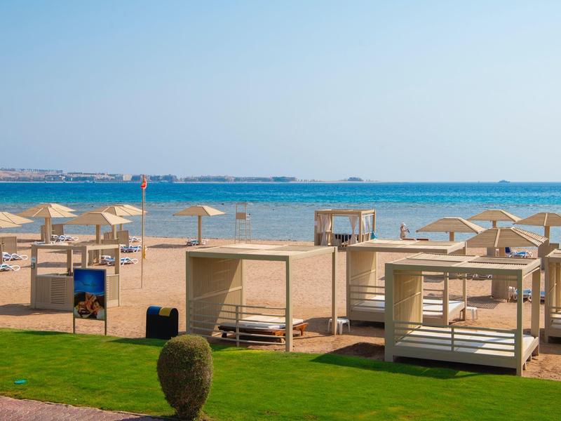 Strand mit weißen Liegen, Holzpavillons, grüner Wiese und blauem Meer unter klarem Himmel.