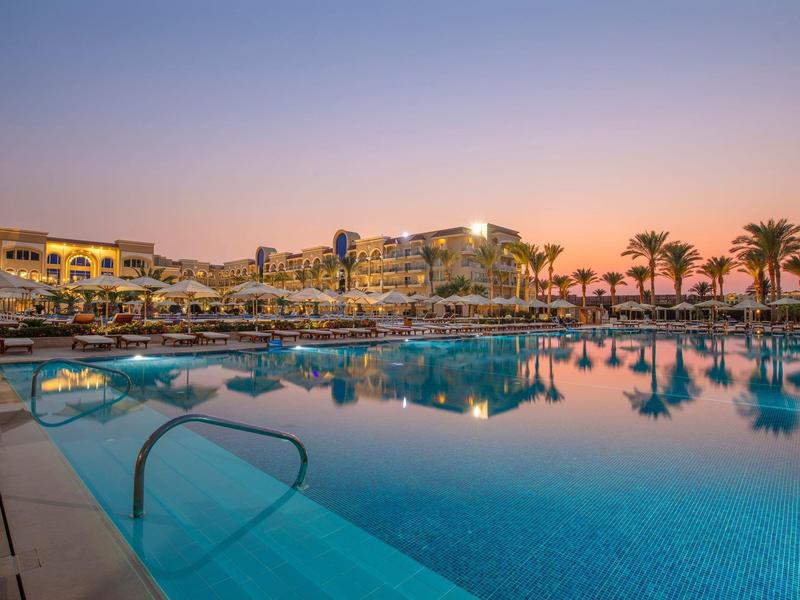 Großer Hotelpool bei Sonnenuntergang mit Liegestühlen, Palmen und Gebäuden im Hintergrund.