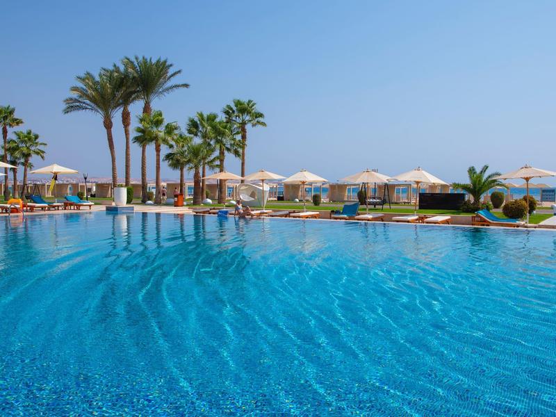 Großer blauer Pool mit Palmen, Sonnenschirmen und Liegestühlen unter hellem Himmel.