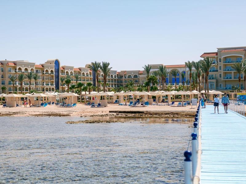 Ein breiter Steg führt über ruhiges Wasser zu einem Strand mit Sonnenschirmen und Palmen, dahinter Hotels.