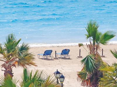 Sandstrand mit zwei leeren blauen Liegestühlen, Palmen und ruhigem, blauem Meer im Hintergrund.