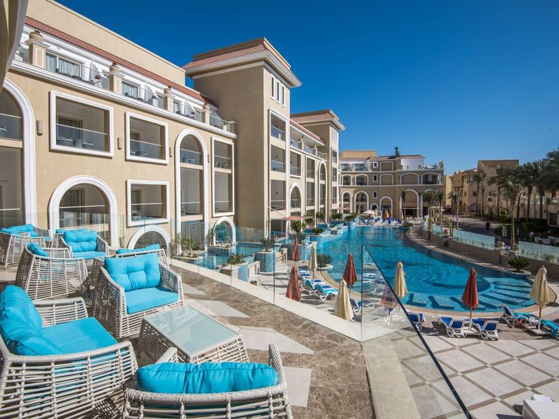 Terrasse d'hôtel avec coussins bleus près d'une grande piscine extérieure et palmiers sous un ciel clair.