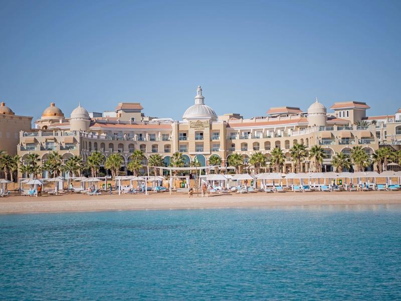 Grand bâtiment hôtelier avec plage de sable et mer bleue au premier plan.