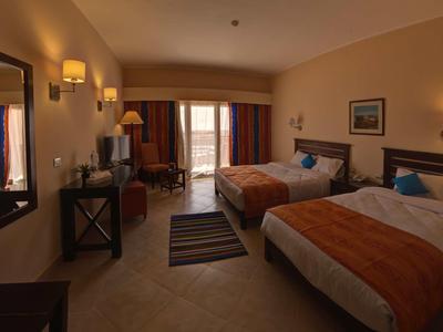 Hotelzimmer mit zwei Betten, orangenen Decken, Teppich, modernen Möbeln und Balkon mit Vorhängen.