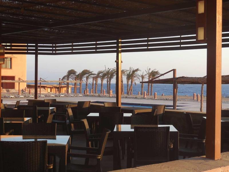 Geräumige Außenterrasse mit vielen Tischen und Stühlen, Blick auf Palmen und das Meer bei Sonnenuntergang.