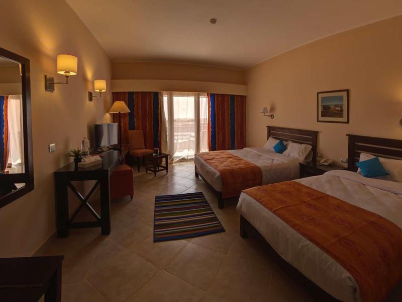 Hotelzimmer mit zwei Betten, orangenen Decken, Teppich, modernen Möbeln und Balkon mit Vorhängen.