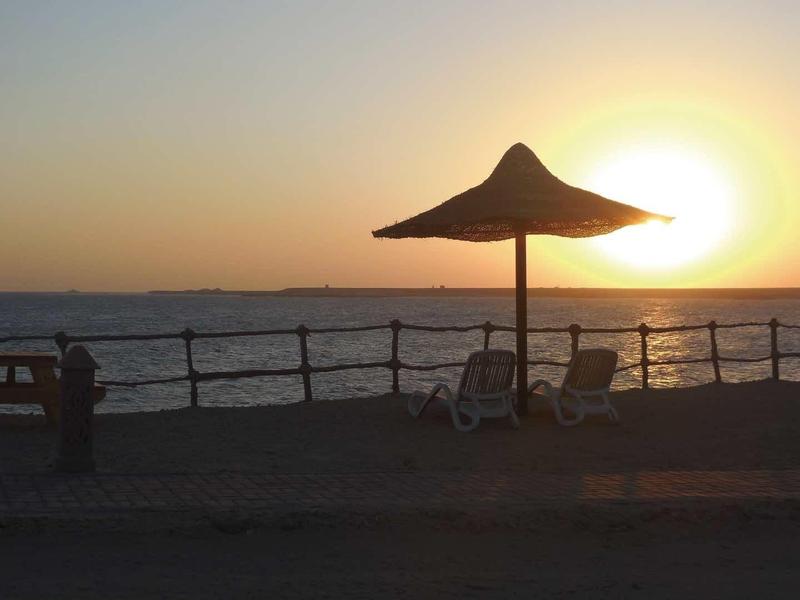 Strand mit Sonnenschirm, Stühlen und Personen, die den Sonnenuntergang über dem Meer beobachten.