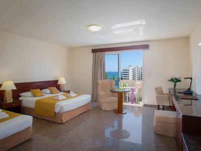 Helles Hotelzimmer mit zwei Betten, Stuhl, Tisch, Schreibtisch, TV und Blick auf Stadtwohnungen.