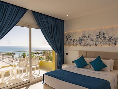 Habitación de hotel luminosa con cama doble, detalles azules y vista al mar desde el balcón.