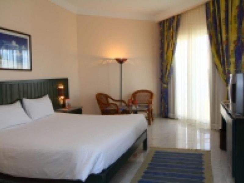 Helles Hotelzimmer mit großem Bett, zwei Stühlen, Stehlampe und Fernseher, Gardinen grün-gelb.