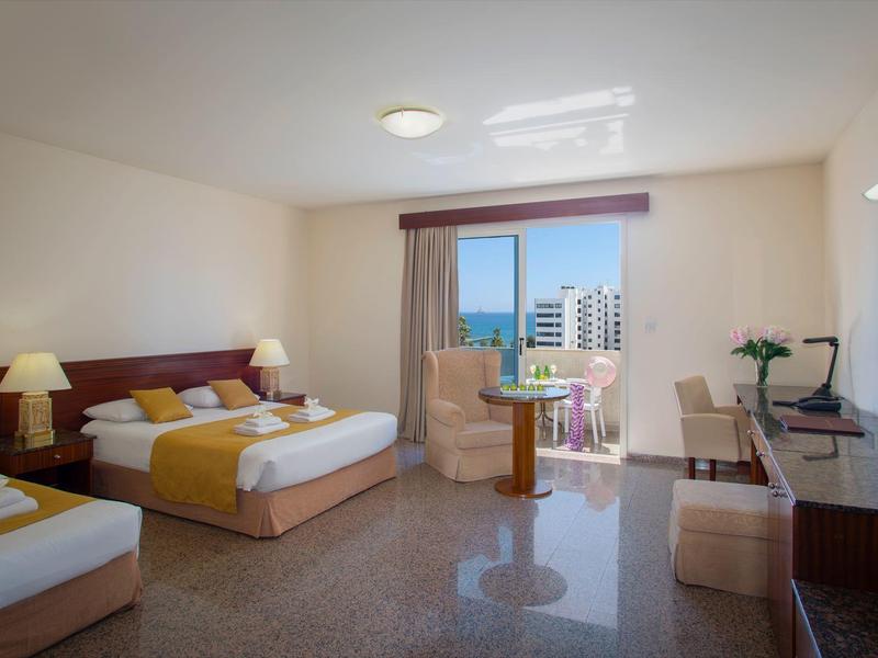 Helles Hotelzimmer mit zwei Betten, Stuhl, Tisch, Schreibtisch, TV und Blick auf Stadtwohnungen.