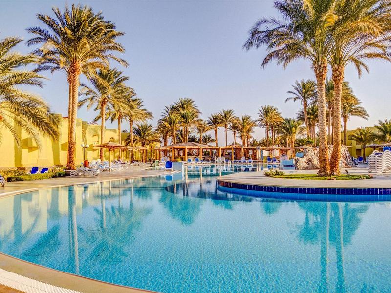 Großer blauer Pool umgeben von Palmen, Liegestühlen und gelben Gebäuden bei klarem Himmel.