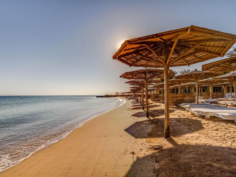 Leerer Sandstrand mit Holzsonnenschirmen und ruhigem, blauem Meer unter klarem Himmel.