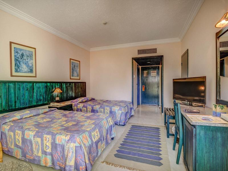 Ein einfaches Hotelzimmer mit zwei Einzelbetten, bunten Bettdecken, grünem Mobiliar und einem Fernseher.