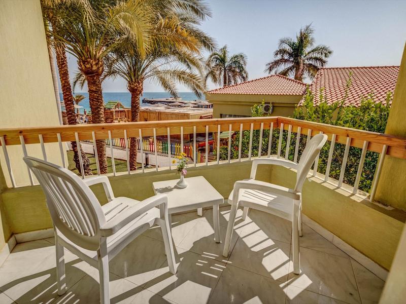 Balkon mit zwei weißen Stühlen und kleinem Tisch, Blick auf Palmen und Meer im Hintergrund.