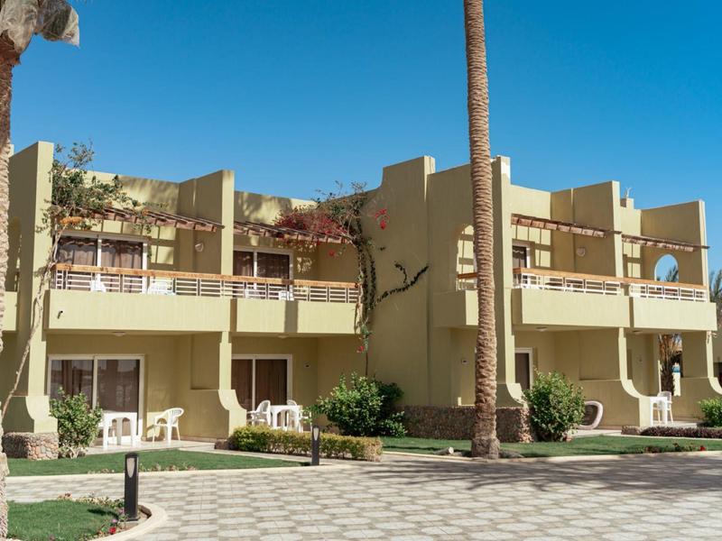 Dvoupodlažní hotelová budova s balkony a palmami pod jasnou oblohou.