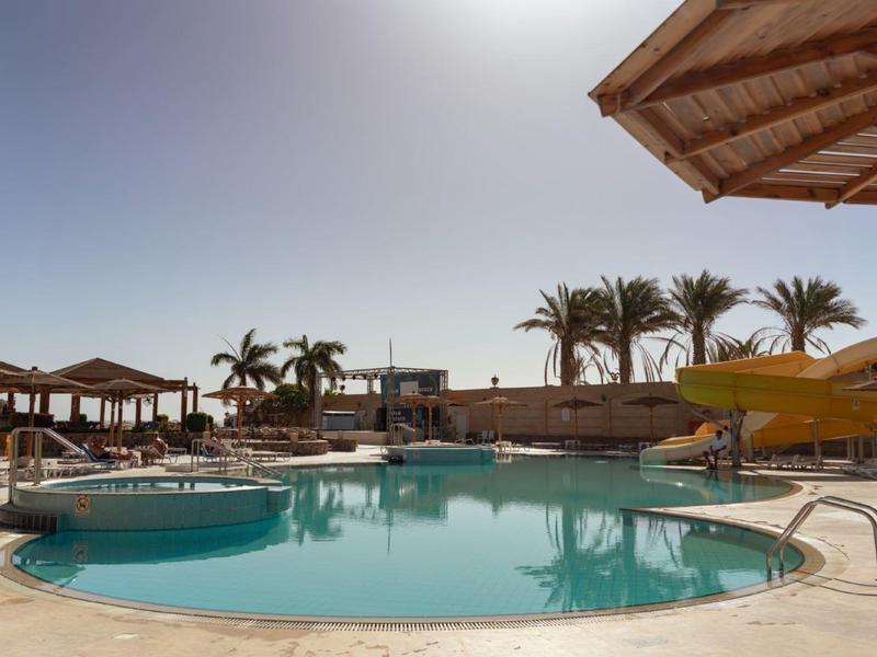 Velký bazén s tobogánem, slunečníky a palmami za slunečného dne.