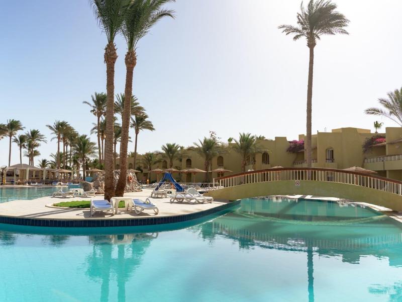 Swimmingpool mit Sonnenliegen, Palmen und Brücke im Hotelresort bei hellem Tageslicht