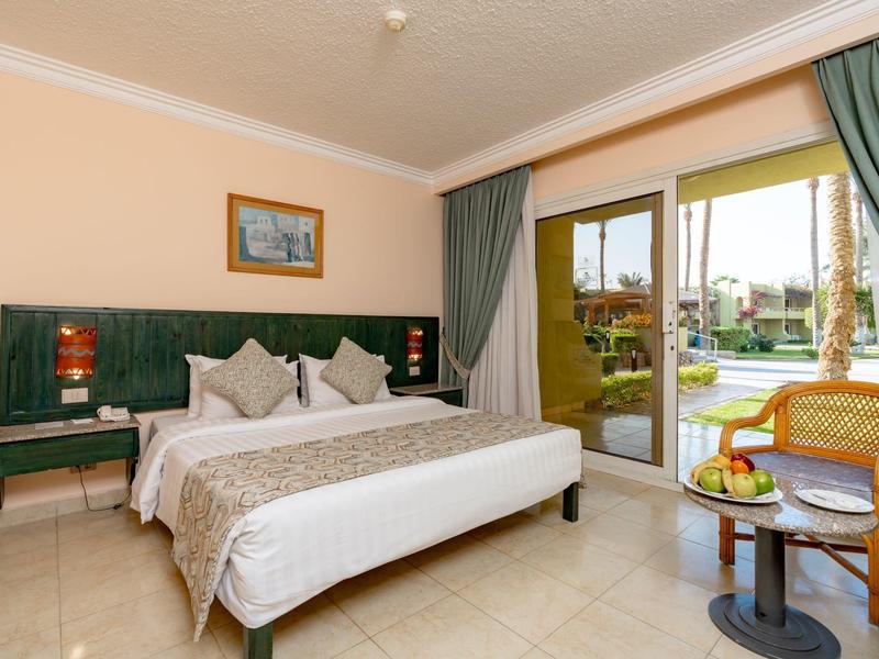 Helles Hotelzimmer mit Doppelbett, Nachttischen, Sitzecke und Blick auf den Pool und Garten.