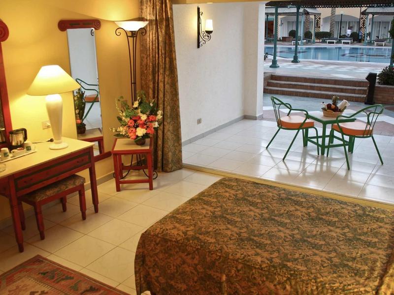 Gemütliches Hotelzimmer mit Bett, Schreibtisch, Spiegel, Stuhl und Terrasse mit Tisch und Stühlen neben Pool.