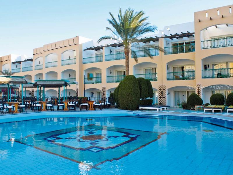 Ein großer, klarer Pool vor einem hellen Hotelgebäude mit Bögen und einer Palme.