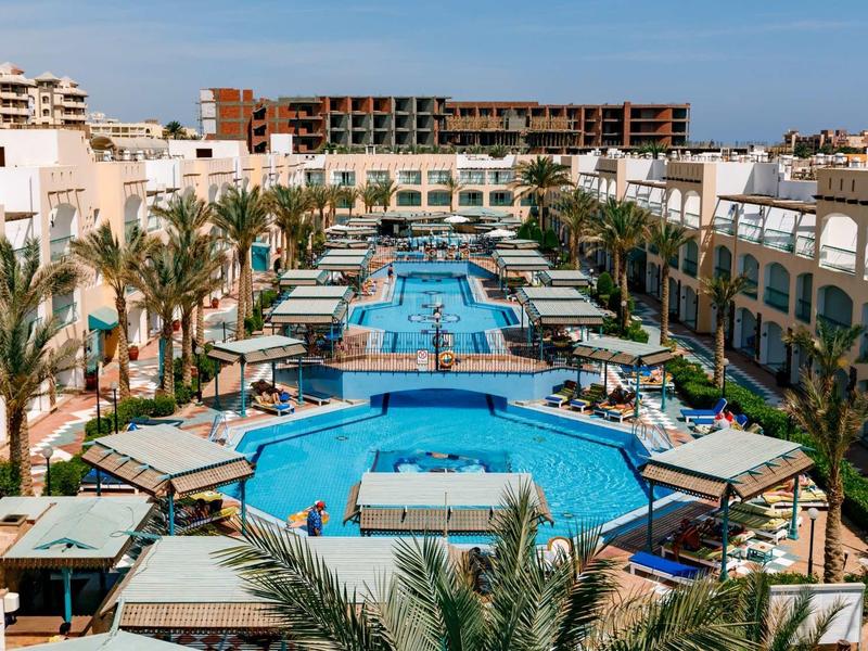 Großer Hotelpool mit mehreren Becken, Palmen, Liegestühlen und weißen Gebäuden unter blauem Himmel.