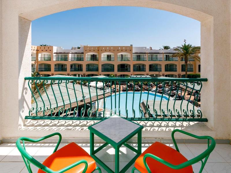 Blick von Balkon mit grünem Geländer, zwei grünen Stühlen mit roten Kissen und Pool vor Gebäude.