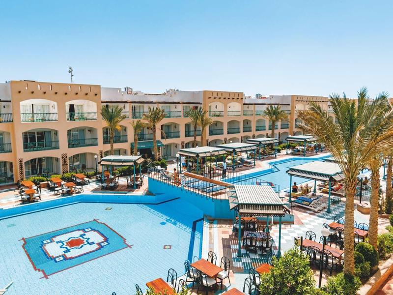Hotel mit großen blauen Pools, Liegestühlen, Palmen und dreistöckigem Gebäude im Hintergrund.