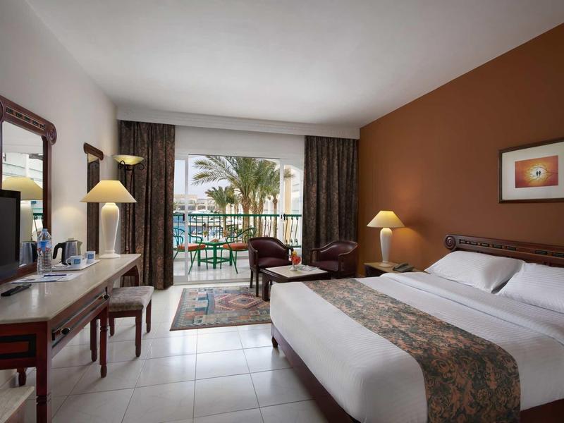 Helles Hotelzimmer mit Doppelbett, brauner Wand, Schreibtisch, TV und Balkon mit Palmenblick.