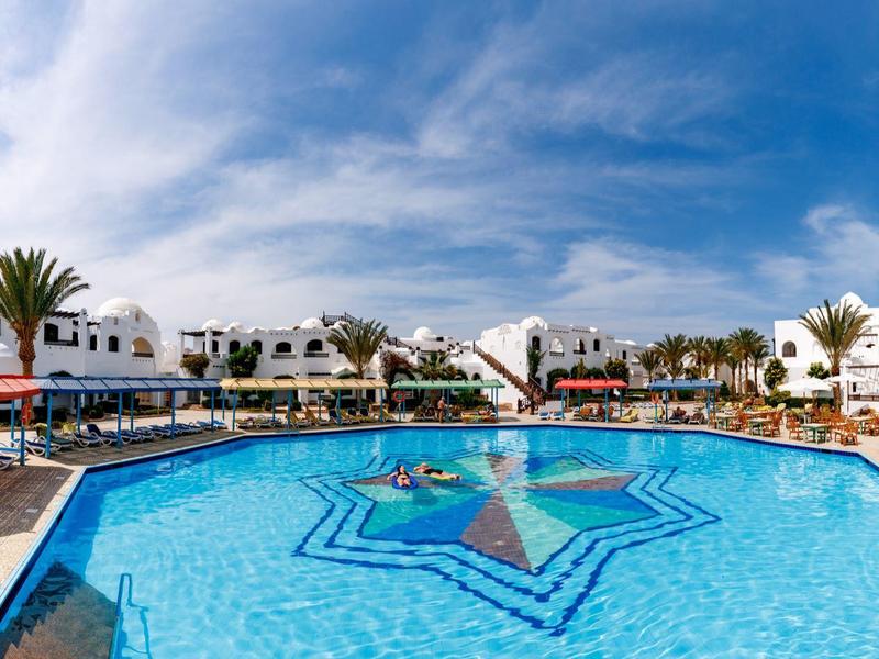 Großer Hotelpool mit Sternmuster, blauem Wasser und umliegenden weißen Gebäuden.