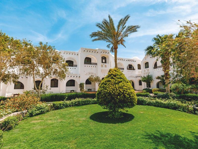 Hotel mit weißer Fassade und gepflegtem Garten mit Palmen und grünem Rasen.