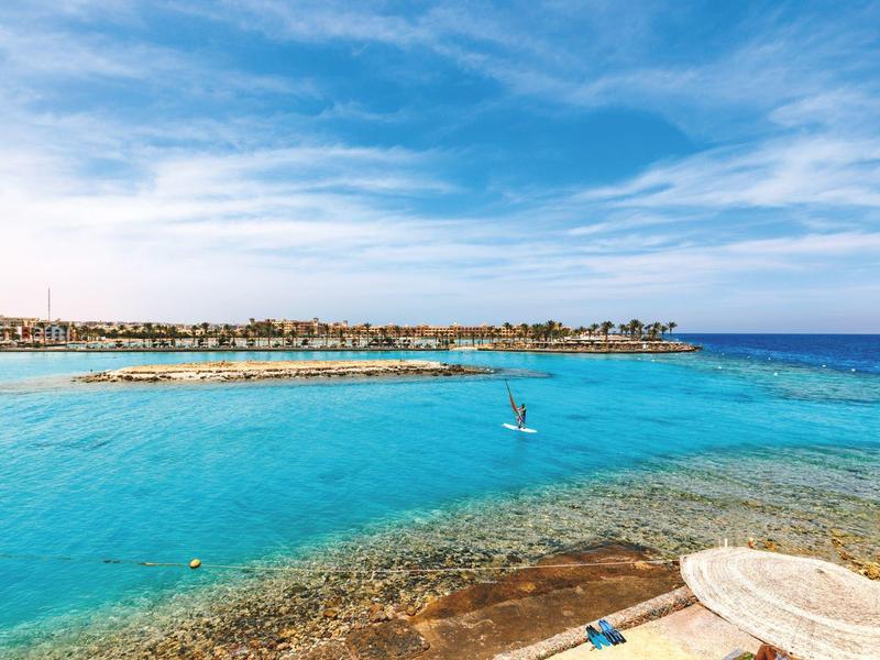 Klares türkisfarbenes Meer mit Booten vor einer flachen Küste unter blauem Himmel.